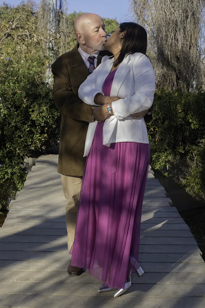 Un paso detrás de Zoila, Carlos se inclina para buscarla, y ella, al sentirlo cerca, gira el rostro hacia atrás como quien responde al llamado más dulce. Sus labios se encuentran en un beso que une dos caminos en uno solo, un gesto suave que parece suspender el mundo.