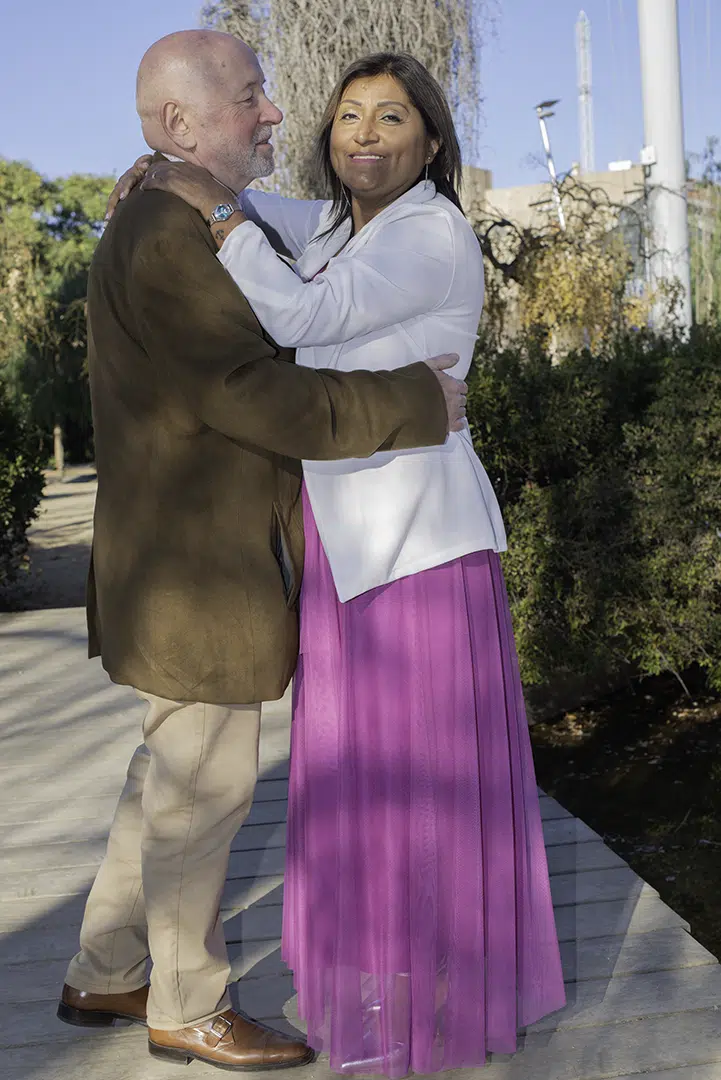 Recién unidos en promesa eterna, Carlos rodea la cintura de Zoila con la ternura de quien ha encontrado su hogar. La contempla con una sonrisa que habla de destino, mientras ella mira a la cámara iluminada por la dicha de un amor recién celebrado. En este instante suspendido, sus miradas cuentan la historia de dos almas que comienzan a latir al mismo ritmo.