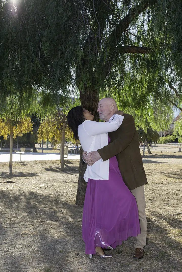 Junto al árbol, Zoila se inclina con gracia, sostenida por un pie, mientras sus ojos se encuentran con los de Carlos; él la abraza firme, como guardando su equilibrio, y en ese instante sus miradas cuentan la fuerza y complicidad de su amor.