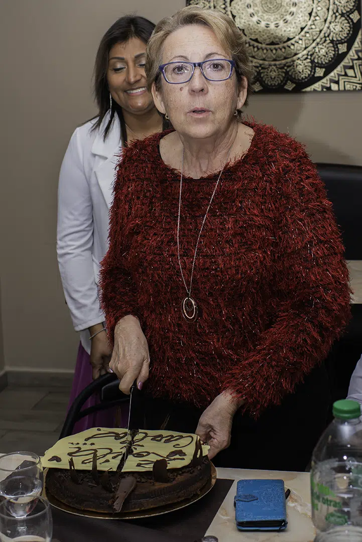 Marta corta el pastel con mirada audaz, mientras Zoila la observa detrás con una sonrisa cómplice, capturando un instante de juego, ternura y alegría compartida.