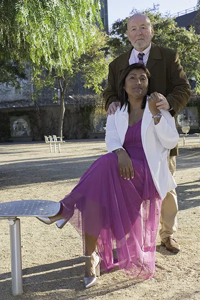 La serenidad del parque los rodea, mientras Carlos, detrás de ella reposa las manos sobre sus hombros en un recordatorio del amor que la sostiene, siempre suave, siempre firme.